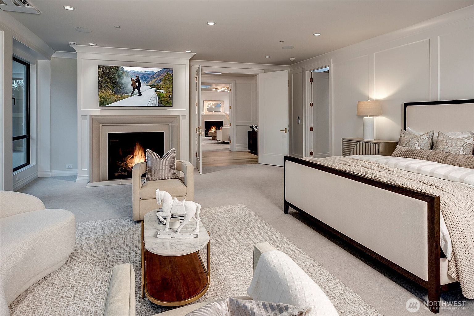 This luxurious primary bedroom features a sophisticated neutral color palette, plush carpeting, and a grand fireplace with an integrated television. The room is anchored by a large, elegant bed with a dark wood frame and is complemented by a cozy seating area with a marble-topped coffee table and a decorative horse sculpture. A wide doorway leads into an adjacent room, creating an open and airy flow that emphasizes the home's spacious and high-end design.