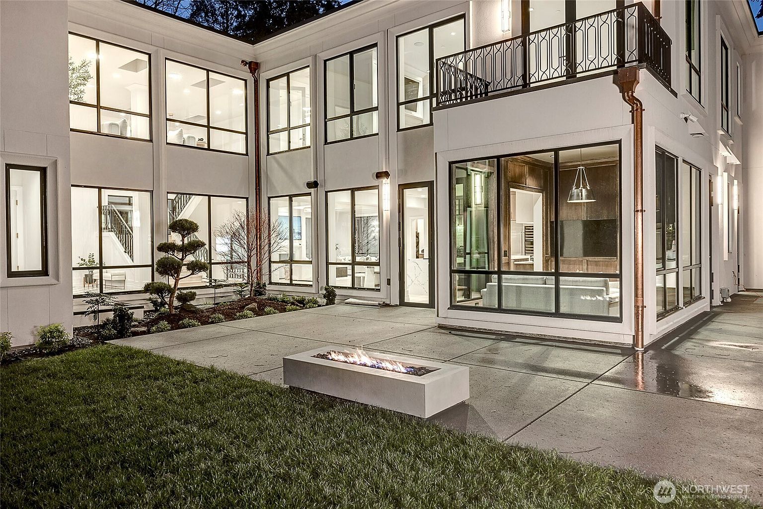 This modern, multi-story luxury home features a striking U-shaped architectural design with expansive floor-to-ceiling windows that create a seamless indoor-outdoor connection. The rear patio includes a sleek, rectangular concrete fire pit set against a backdrop of manicured landscaping and a lush green lawn. The evening lighting highlights the home's contemporary aesthetic, emphasizing the clean lines and sophisticated outdoor living space.