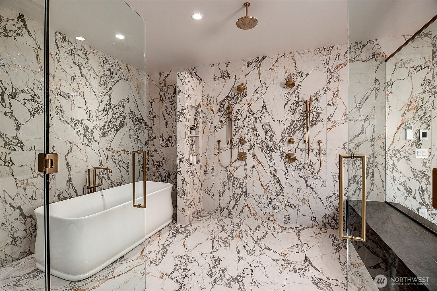 This luxurious primary bathroom features a wet room design with floor-to-ceiling marble tile, a freestanding soaking tub, and a spacious walk-in shower with dual showerheads. The space is accented with elegant gold-toned fixtures and hardware, creating a sophisticated and high-end aesthetic. The perspective is a wide-angle shot that captures the seamless integration of the bathing and showering areas behind a glass enclosure.