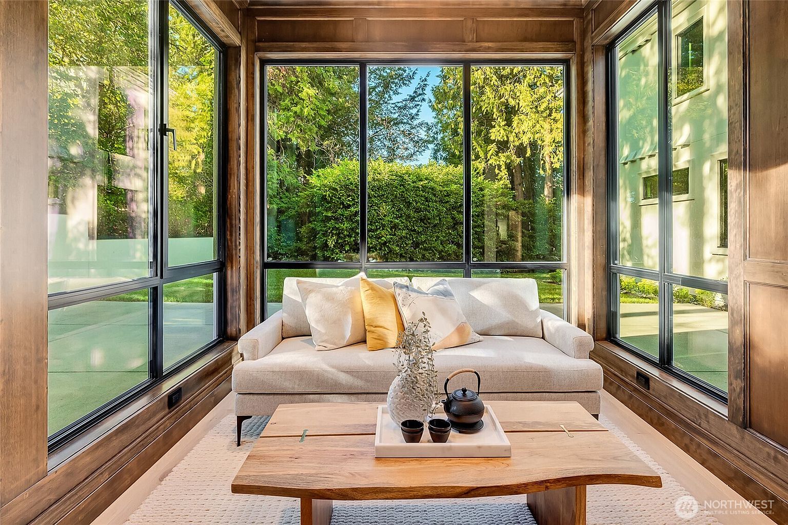 This elegant sunroom features floor-to-ceiling windows that frame a lush, green outdoor view, creating a seamless connection with nature. The space is anchored by a comfortable, light-toned sofa adorned with decorative pillows and a rustic, live-edge wood coffee table topped with a tea set. Rich wood paneling on the walls adds warmth and sophistication to this serene, light-filled retreat.