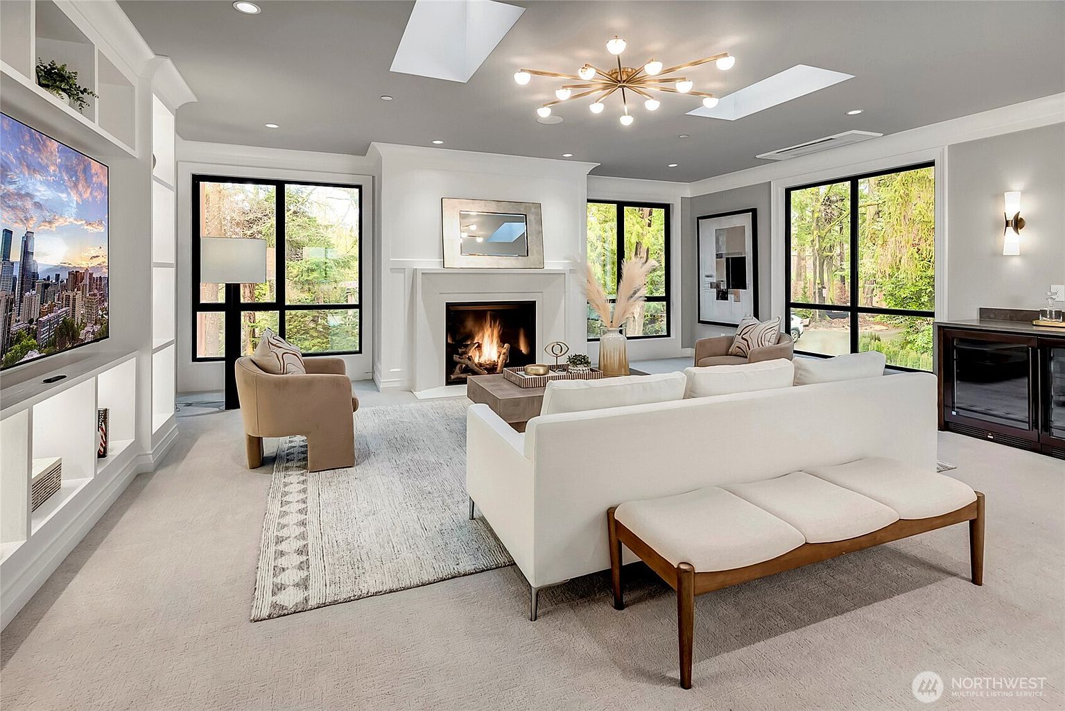 This sophisticated living room features a bright, open-concept design with a central fireplace, a large white sofa, and a matching bench, all anchored by a textured area rug. The space is illuminated by a modern sputnik-style chandelier and natural light streaming through large windows and skylights, while built-in shelving provides a sleek storage solution. The neutral color palette and clean lines create an inviting, high-end atmosphere perfect for relaxation or entertaining.