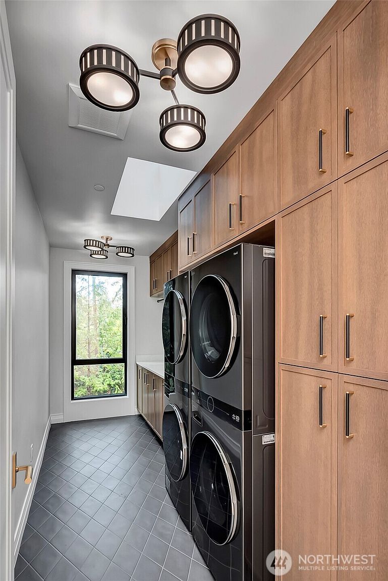 This modern, high-end laundry room features sleek, dark-toned stacked washer and dryer units set against warm, light-wood cabinetry that provides ample storage. The space is illuminated by unique, contemporary multi-light fixtures and natural light from a tall window, all set above a sophisticated dark-tiled floor laid on a diagonal. The overall aesthetic is clean, organized, and luxurious, offering a functional yet stylish utility space.