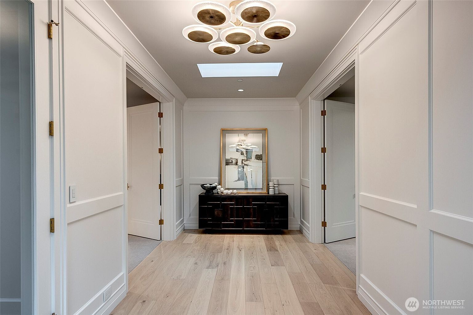 This elegant hallway features light-toned hardwood flooring and crisp white wainscoting, creating a bright and sophisticated atmosphere. A striking, multi-tiered circular chandelier serves as a focal point above, while a dark wood console table adorned with art and decor anchors the end of the corridor. The perspective is a centered, symmetrical shot that emphasizes the clean lines and high-end architectural details of the home.