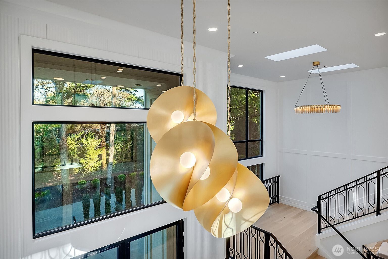 This high-angle shot captures a dramatic, multi-story entryway featuring a striking, sculptural gold pendant light fixture as the focal point. The space is characterized by clean white walls, large black-framed windows that offer a view of the outdoors, and a sophisticated staircase with a black iron railing. The overall aesthetic is modern, luxurious, and airy, emphasizing the home's vertical scale and high-end design details.