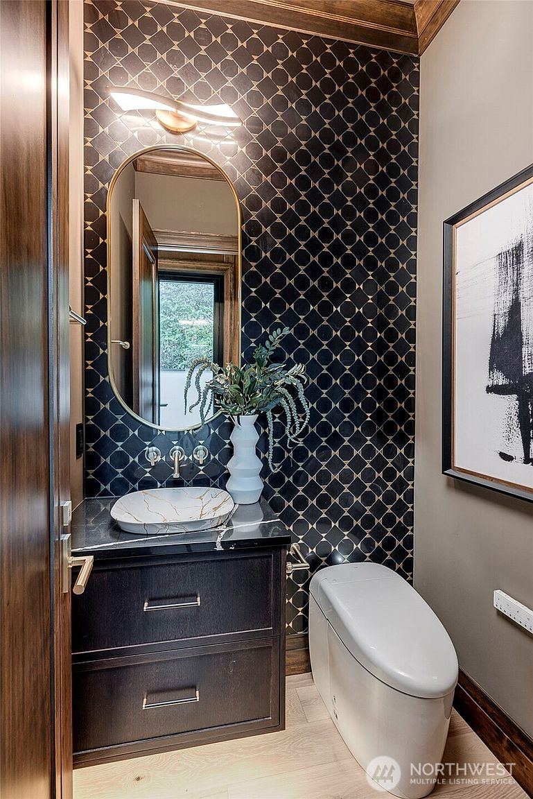 This sophisticated powder room features a striking black and gold geometric patterned tile accent wall that serves as a dramatic focal point. The space is equipped with a sleek, dark wood vanity topped with a marble vessel sink, a modern arched mirror, and a minimalist white toilet. The combination of warm wood tones, bold patterns, and elegant lighting creates a luxurious and contemporary atmosphere.