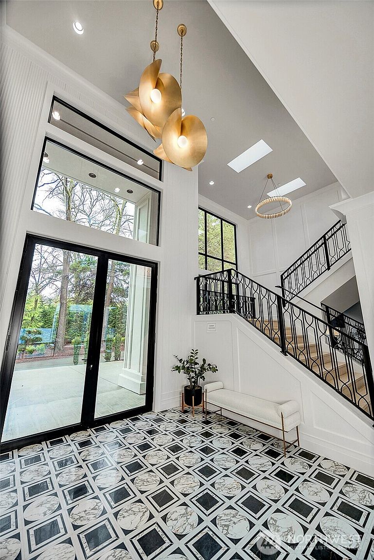 This grand, double-height entryway features a striking black-and-white geometric marble floor and a dramatic gold sculptural chandelier that serves as a focal point. A sleek white bench sits beneath a staircase with intricate black iron railings, while expansive floor-to-ceiling windows flood the space with natural light. The overall aesthetic is one of sophisticated, modern luxury with a clean, high-contrast color palette.