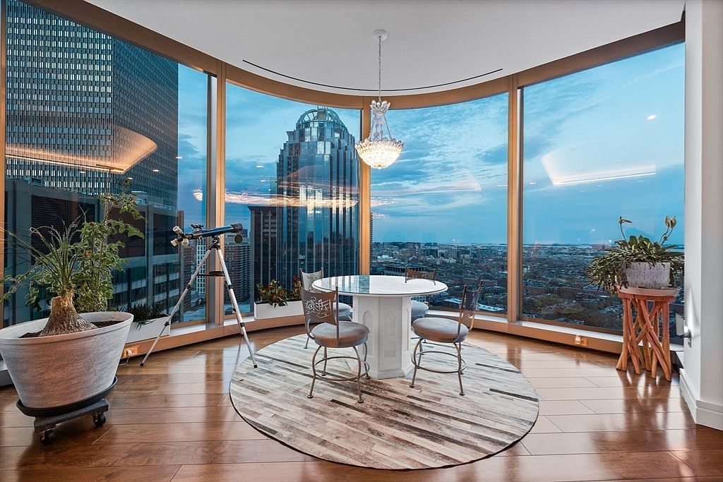 This is an interior shot of a dining room in a high-rise apartment, featuring a round table with chairs set on a circular rug. The room is surrounded by curved floor-to-ceiling windows offering panoramic city views, with a telescope positioned to take advantage of the scenery. A chandelier hangs above the table, adding a touch of elegance to the modern space.