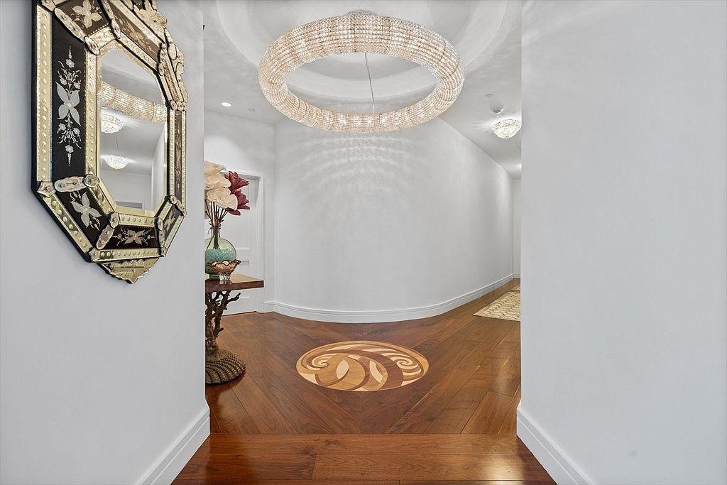 This interior shot showcases an elegant hallway with a striking circular chandelier as a focal point. The hardwood flooring features a decorative inlay, adding a touch of sophistication. A decorative mirror hangs on the wall to the left, and the walls are painted in a neutral tone, creating a bright and inviting space.