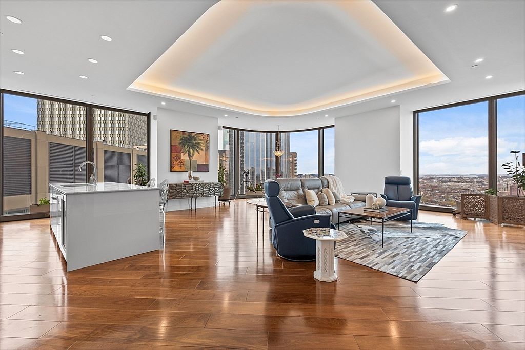 This is a spacious and luxurious living room with floor-to-ceiling windows offering panoramic city views. The room features hardwood floors, a modern ceiling design with recessed lighting, and stylish furniture including a leather sofa and armchairs. A contemporary rug anchors the seating area, creating a comfortable and elegant space.