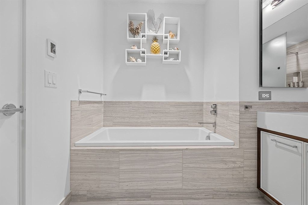 This is a well-lit bathroom featuring a built-in bathtub with light gray stone-like tiling surrounding it. Above the tub, there are decorative white shelves with beach-themed decor, including a gold pineapple. To the right of the tub is a modern sink and vanity, and a mirror is visible above the sink.