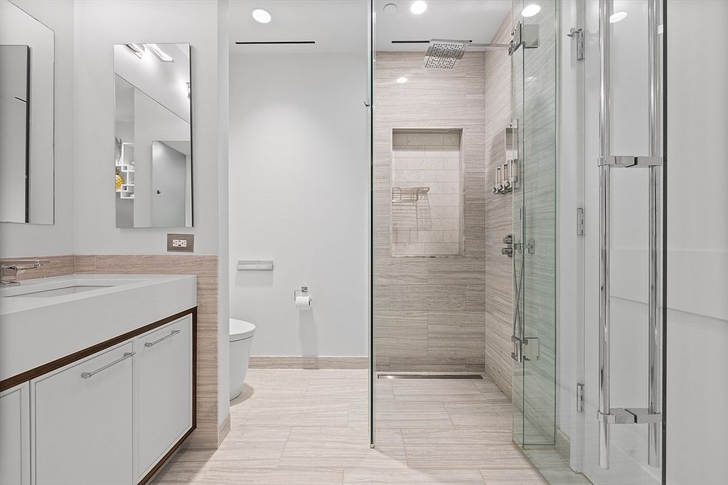 This is a modern primary bathroom featuring a sleek vanity with a white countertop and dark wood trim, a large mirror, and a glass-enclosed shower. The shower has a rainfall showerhead and a built-in niche for toiletries. The floor is tiled with light-colored planks, and the overall aesthetic is clean and minimalist.