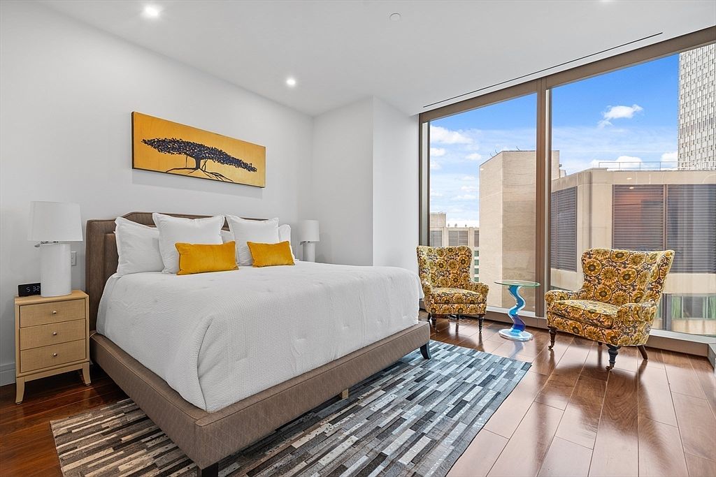 This is a primary bedroom featuring a large bed with white linens and yellow accent pillows, flanked by a wooden nightstand and lamp. The room has large windows offering city views, and two patterned armchairs are positioned near the window. A modern rug adds texture to the hardwood floors, and a piece of artwork hangs above the bed.