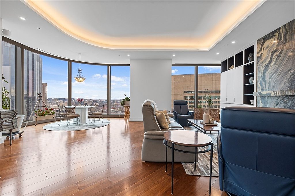This is a luxurious living room featuring hardwood floors, floor-to-ceiling windows with city views, and modern furnishings. The room includes a dining area with a chandelier, a seating area with a sofa and armchairs, and a built-in shelving unit with a marble accent wall. The overall impression is one of elegance and sophistication.
