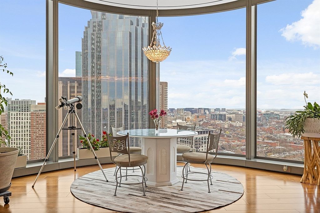 This is an interior shot of a dining area in a high-rise apartment, featuring a round glass-top table with four chairs set on a circular rug. A chandelier hangs above the table, and a telescope stands near the curved floor-to-ceiling windows, offering a panoramic city view. The room exudes a modern and luxurious ambiance.