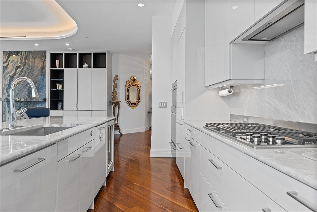 This is a modern kitchen with sleek, white cabinetry and stainless steel appliances. The kitchen features a large island with a marble countertop and a built-in wine cooler. The hardwood floors add warmth to the space, and the overall design is clean and contemporary, creating a luxurious and functional cooking area.
