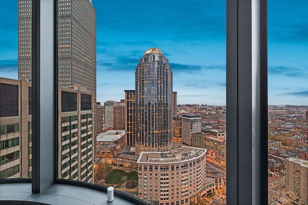 This image showcases a stunning cityscape view from what appears to be a high-rise apartment or condominium. The view is framed by the window structure, offering a panoramic perspective of the surrounding buildings and urban landscape. The interior space is suggested by the window frame and a small white object on the window sill, emphasizing the luxurious and modern living experience.
