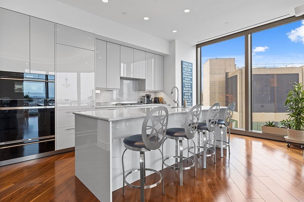 This is a modern kitchen with sleek, glossy white cabinetry and a large island with seating. Stainless steel appliances are integrated into the design, and a large window provides natural light and a city view. The hardwood floors add warmth to the contemporary space.