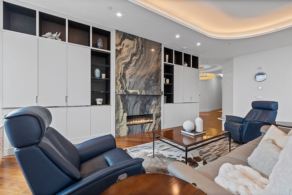 This is an interior shot of a luxurious living room featuring a modern fireplace with a striking marble surround, flanked by custom built-in cabinetry with open shelving. The room is furnished with comfortable leather armchairs, a stylish coffee table, and a plush sofa, creating an inviting and sophisticated atmosphere. The hardwood flooring and recessed lighting add to the room's elegance.