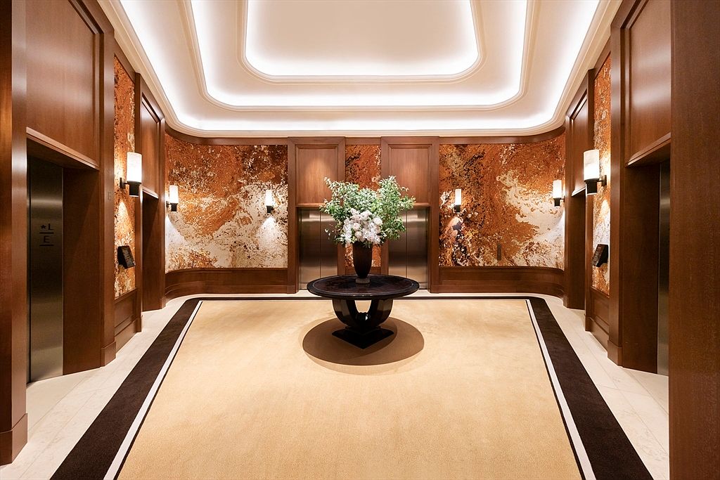 This is an interior shot of an elegant hallway or lobby area, featuring wood-paneled walls and decorative marble accents. Two elevator doors are visible, flanking a central table adorned with a floral arrangement. The space is well-lit with recessed lighting and wall sconces, creating a luxurious and inviting atmosphere.
