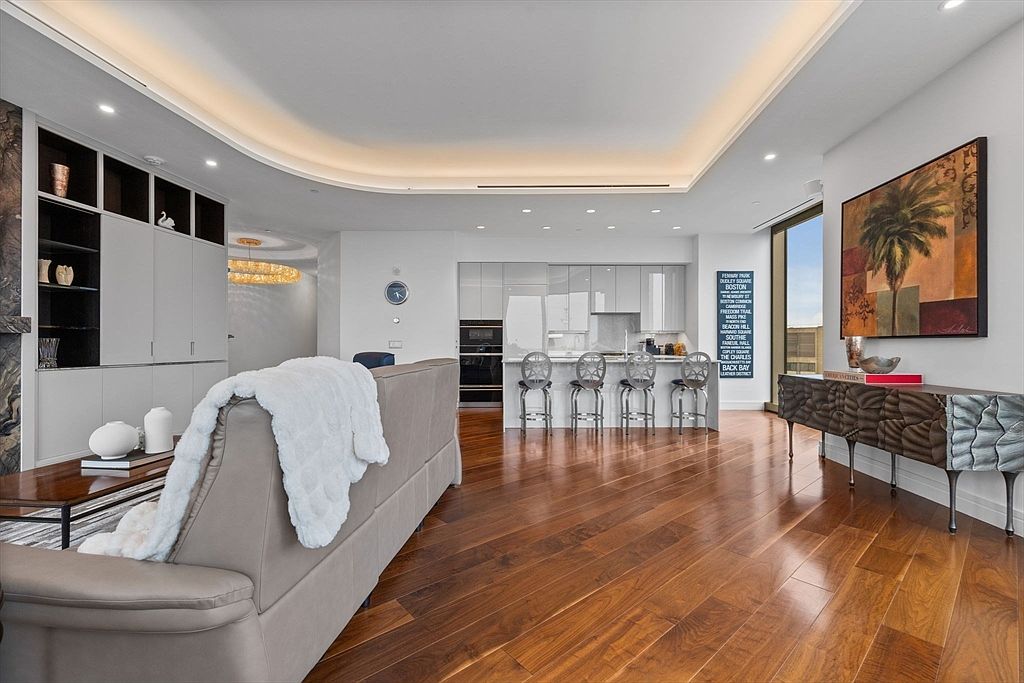 This interior shot showcases a modern living room that seamlessly blends into a kitchen area. The living room features a comfortable sofa with a throw blanket, a coffee table, and a built-in shelving unit. The kitchen boasts sleek, minimalist cabinetry and bar seating, all under a contemporary lighting design, creating an open and inviting space.
