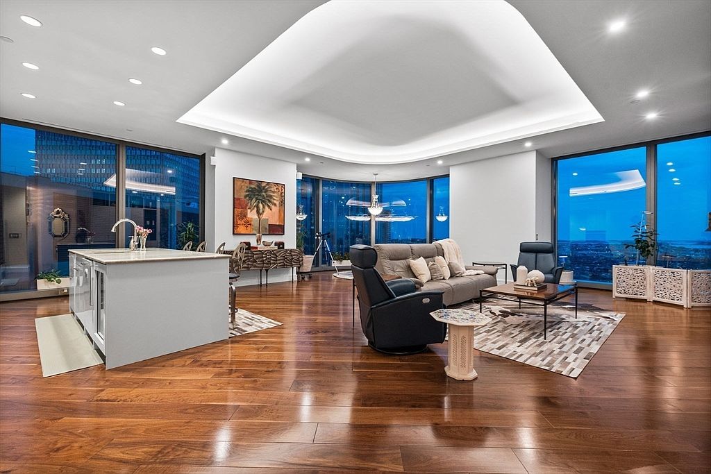 This is a luxurious living room featuring hardwood floors, floor-to-ceiling windows with city views, and a modern design. The room includes a gray sofa, a black recliner, a coffee table, and a kitchen island, all arranged on a patterned rug. The space is well-lit with recessed lighting and a unique ceiling design, creating an open and inviting atmosphere.