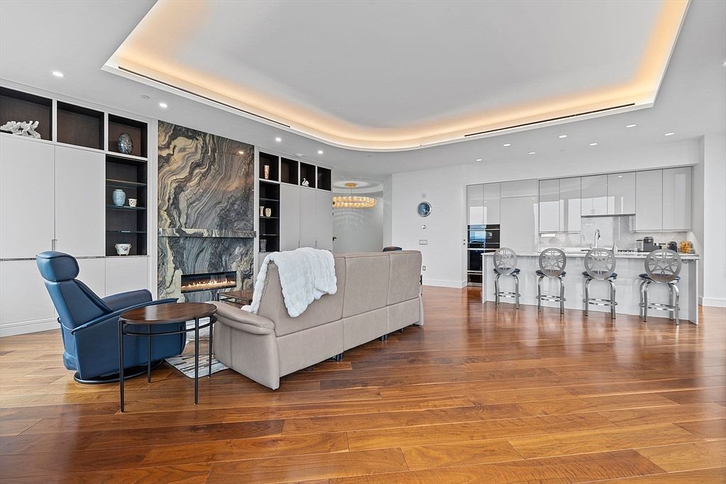 This is a spacious and modern living room featuring hardwood floors, a fireplace with a marble surround, and built-in shelving. The room is furnished with a beige sofa, a blue armchair, and a round side table. The open floor plan seamlessly connects the living area to the kitchen, creating a bright and airy atmosphere.