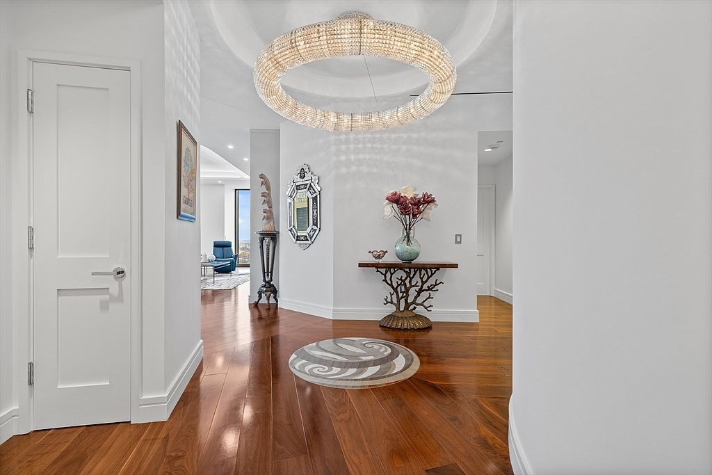 This interior shot showcases a luxurious hallway with rich hardwood flooring and a striking circular chandelier. A decorative rug adds visual interest, while elegant console tables and artwork enhance the sophisticated ambiance. The space is well-lit and exudes a sense of refined elegance.