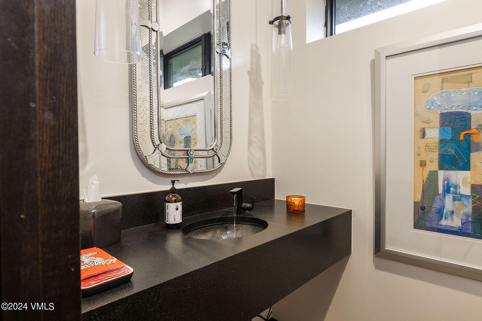 This is a well-appointed guest bathroom featuring a dark countertop with an integrated sink and a modern faucet. A decorative mirror hangs above the sink, complemented by stylish pendant lighting. The walls are painted in a neutral tone, and framed artwork adds a touch of sophistication to the space.