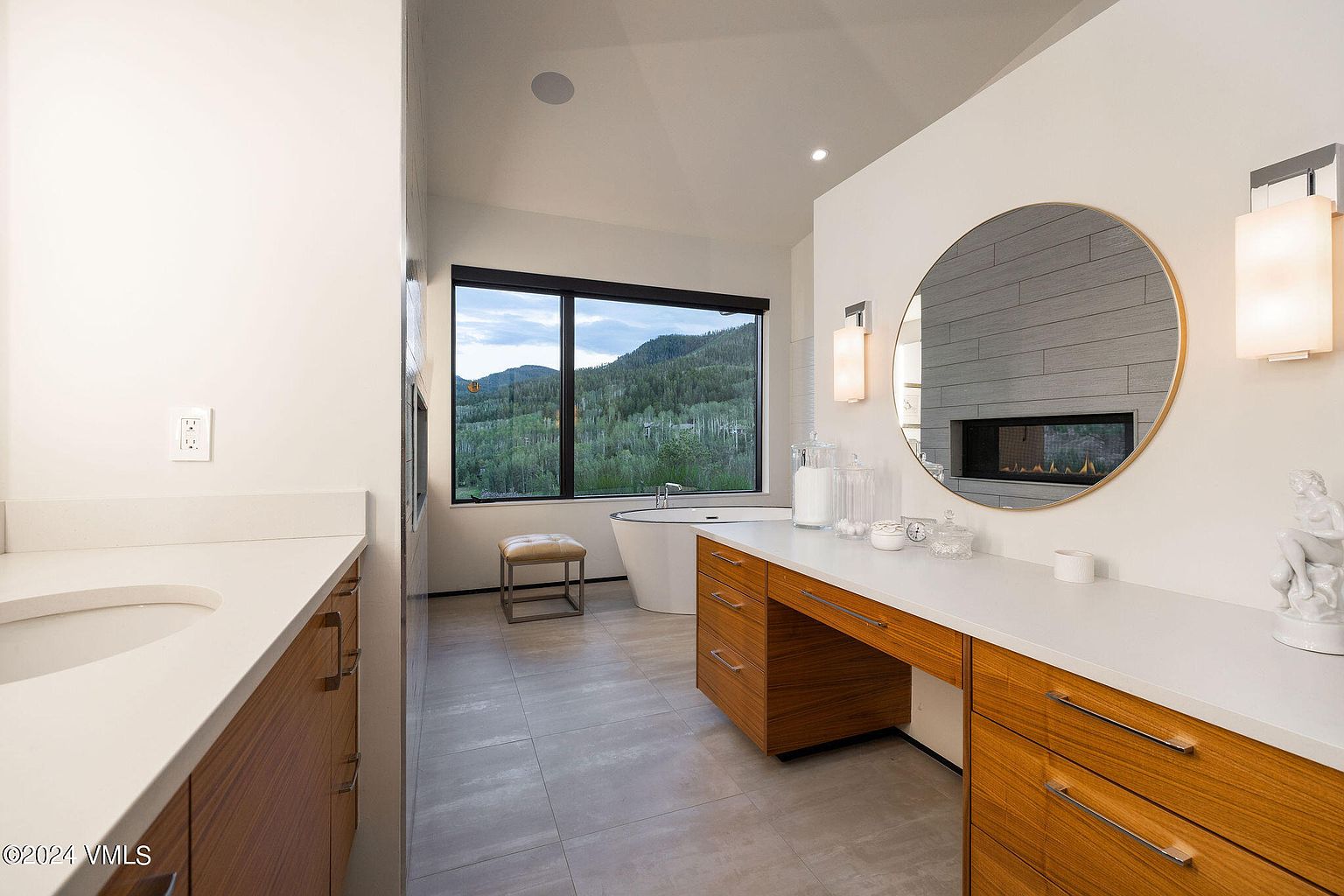 This is a luxurious primary bathroom featuring a modern design with a large window offering scenic mountain views. The bathroom includes a freestanding bathtub, a vanity with wooden cabinets and a white countertop, a round mirror, and a fireplace. The overall impression is one of tranquility and high-end comfort.