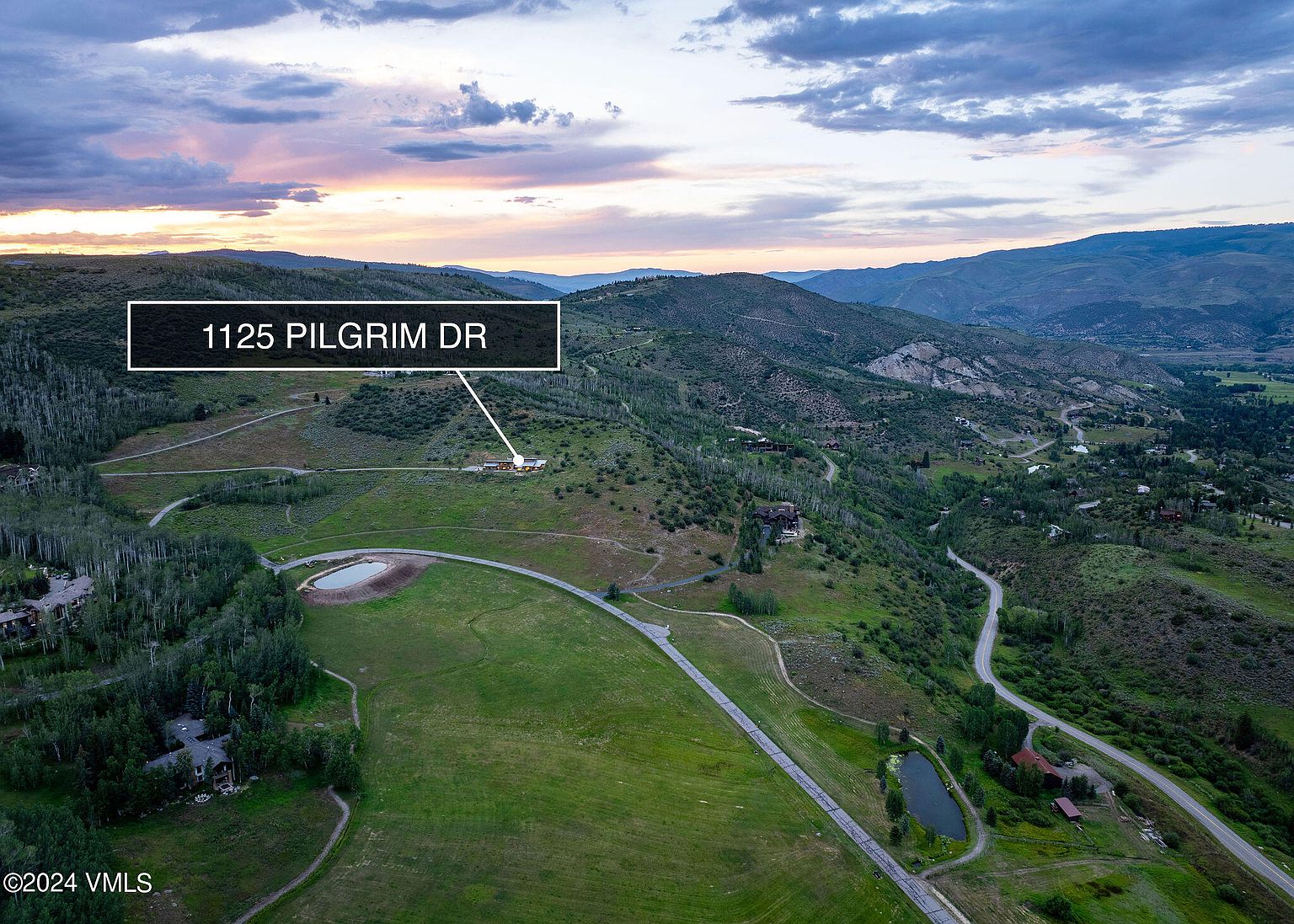 This aerial view showcases a sprawling property nestled in a mountainous landscape, highlighting the home's secluded location and expansive grounds. The property features a large lawn, a pond, and a winding driveway, emphasizing privacy and natural beauty. The surrounding hills and valleys add to the sense of tranquility and exclusivity.