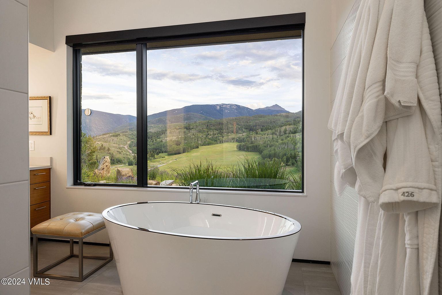 This primary bathroom features a freestanding soaking tub positioned in front of a large window with mountain views. A modern aesthetic is achieved through clean lines, neutral tones, and minimalist fixtures. A plush white robe hangs nearby, adding a touch of luxury.