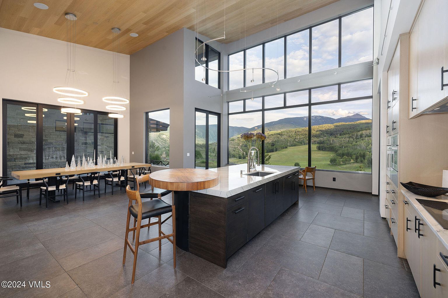 This is a spacious, modern kitchen with a large island featuring a wooden countertop extension and dark cabinetry. Floor-to-ceiling windows offer stunning views of the surrounding landscape, and the open floor plan includes a dining area with a long table. The kitchen is well-lit with modern lighting fixtures and features high-end appliances.
