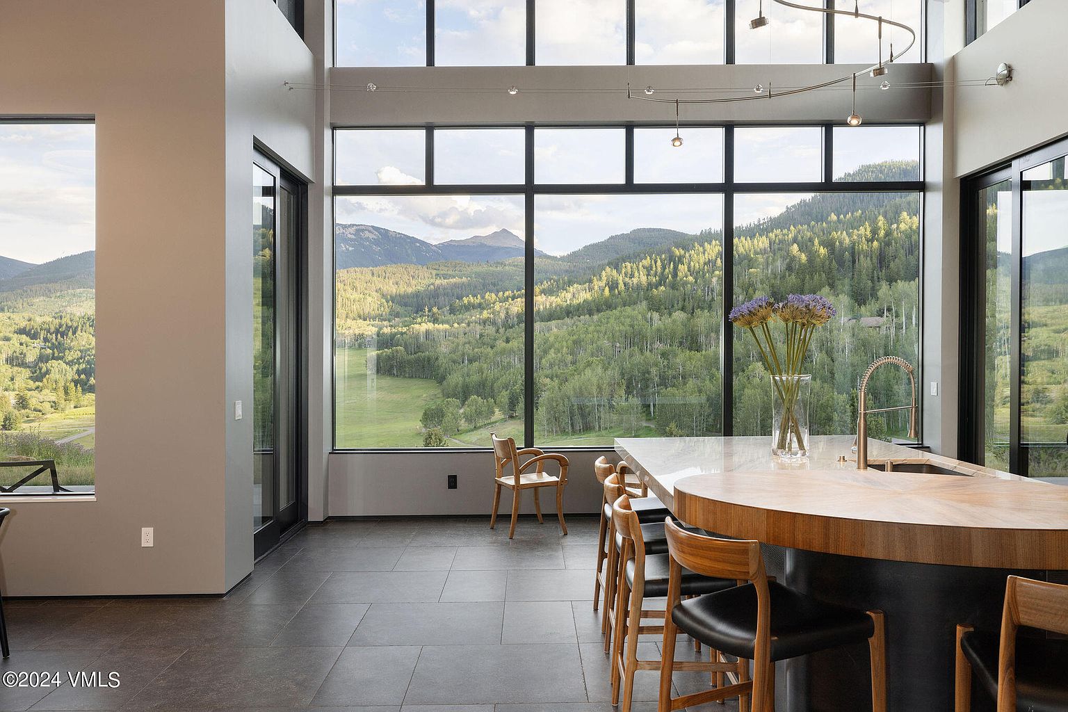 This modern kitchen boasts a large island with a unique countertop and wooden bar stools, complemented by expansive windows offering stunning mountain views. The open floor plan and minimalist design create a bright and inviting space, perfect for both cooking and entertaining. The dark tile flooring adds a touch of sophistication.