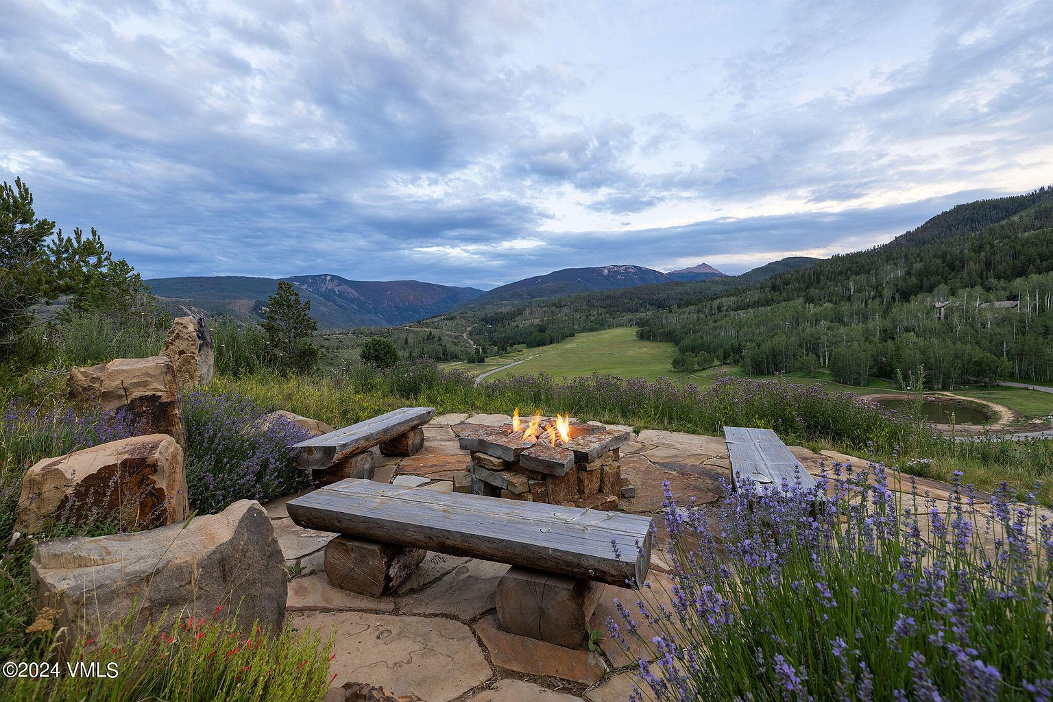 This image showcases a beautifully landscaped yard with a stone fire pit and wooden benches, creating an inviting outdoor gathering space. The surrounding landscape features rolling hills, trees, and a serene mountain backdrop, enhancing the property's appeal. Lavender and other plants add color and texture to the foreground.