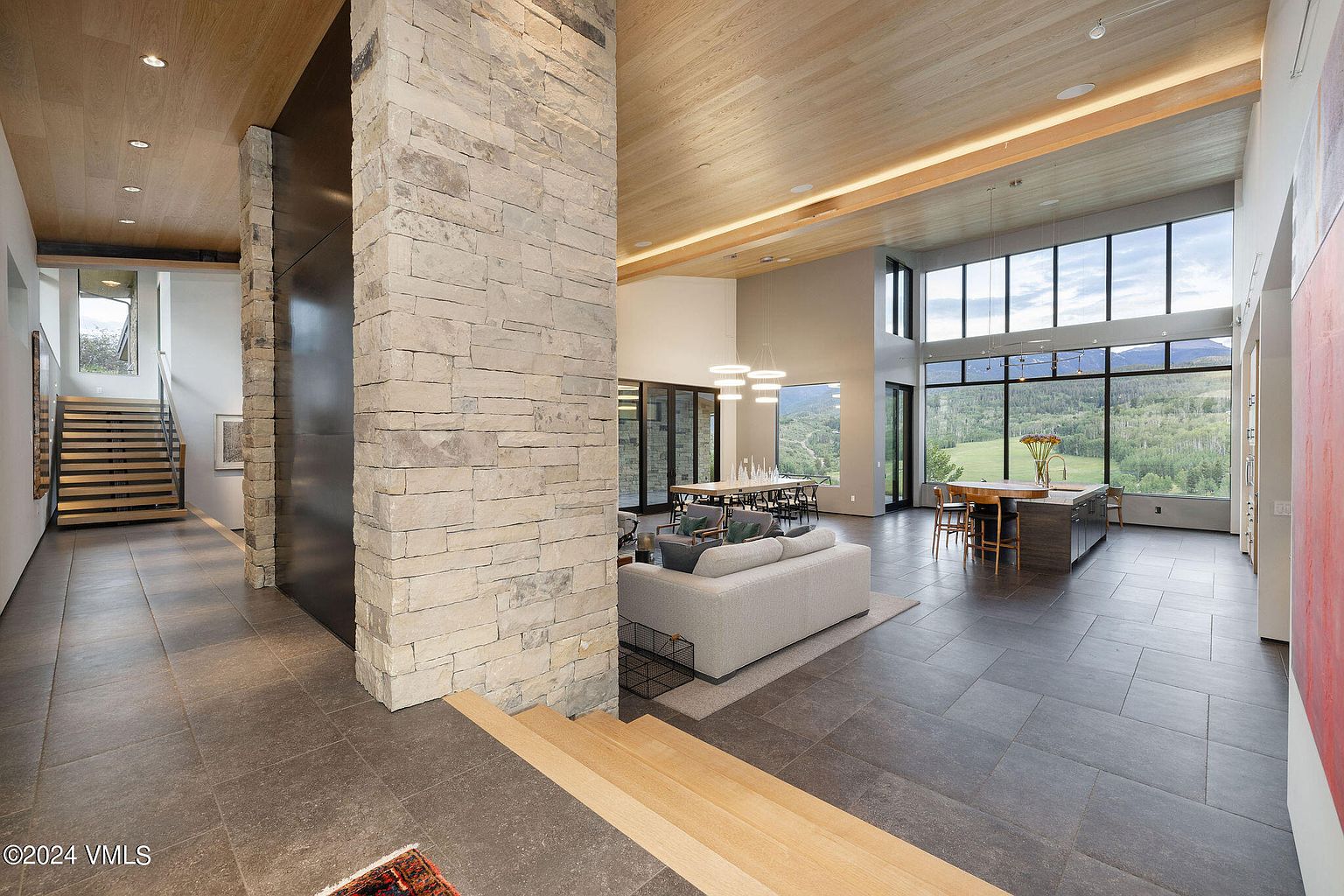 This image showcases a spacious, modern living room with an open floor plan. The room features a stone accent wall, large windows offering scenic views, and contemporary furnishings. The hardwood floors and high ceilings contribute to the luxurious and airy feel of the space, making it an ideal area for relaxation and entertainment.