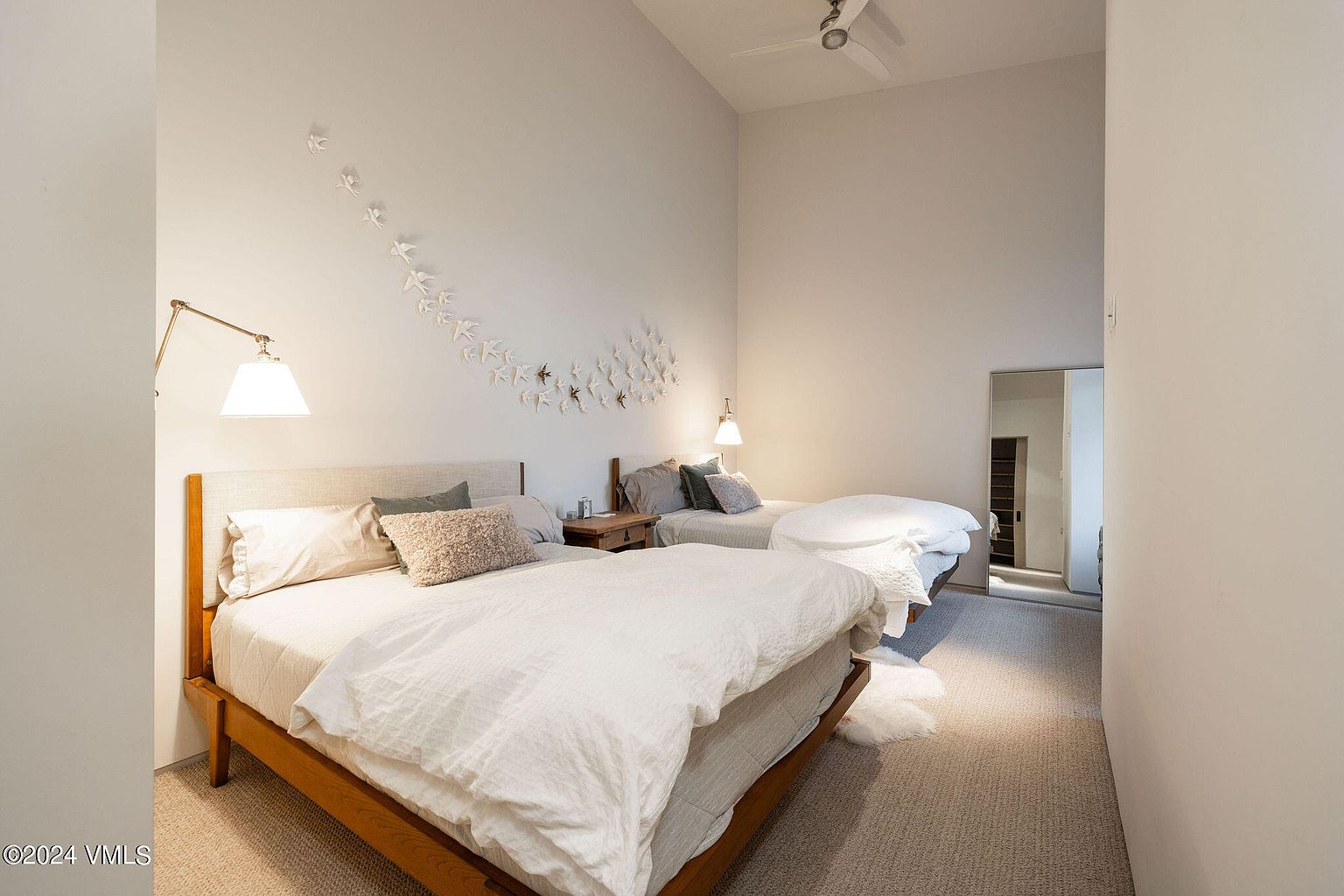 This is a bright and airy guest bedroom featuring two twin beds with light-colored bedding and wooden frames. The room has a minimalist design with white walls, a unique bird-themed wall decoration, and a full-length mirror. The overall impression is clean, comfortable, and inviting.