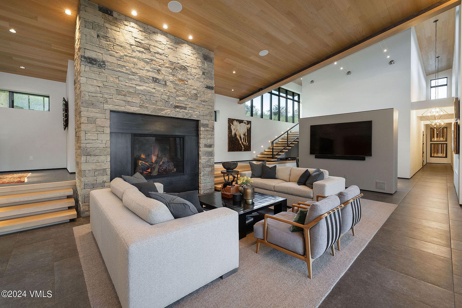 This is a modern living room featuring a stone fireplace, hardwood floors, and contemporary furniture. The room has a high ceiling with recessed lighting and large windows that provide natural light. The overall impression is luxurious and inviting.