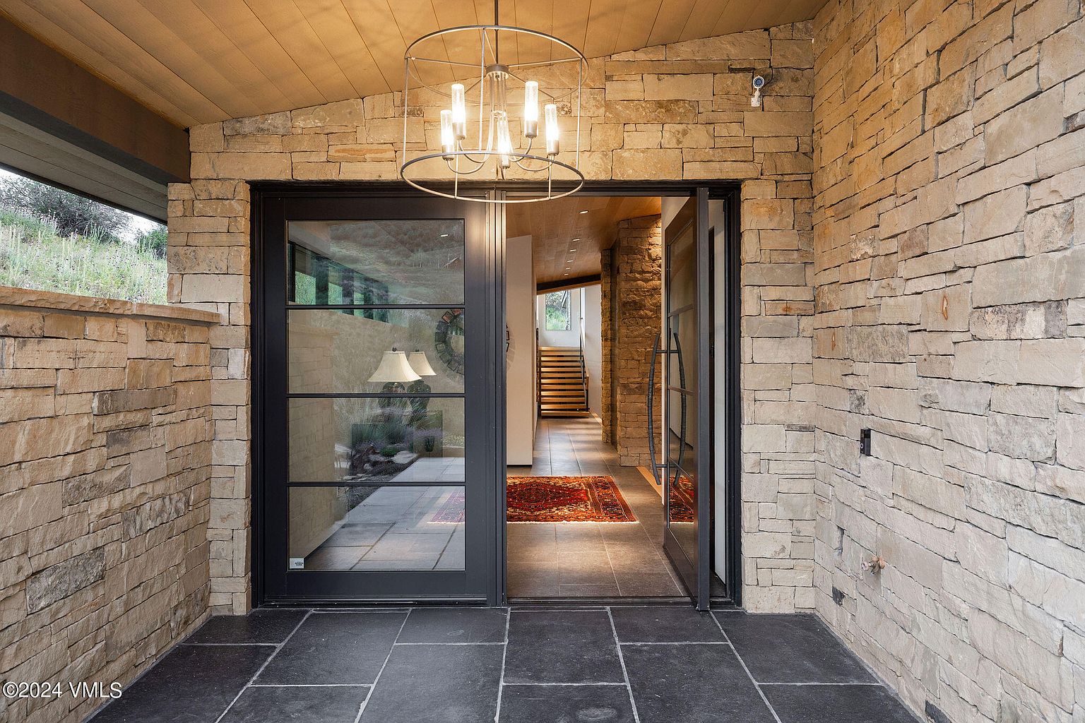 This image showcases a grand entryway with stone walls and black-framed glass doors. A modern chandelier hangs from the wooden ceiling, illuminating the space. The entryway leads into a hallway with a staircase, creating a welcoming and luxurious first impression.