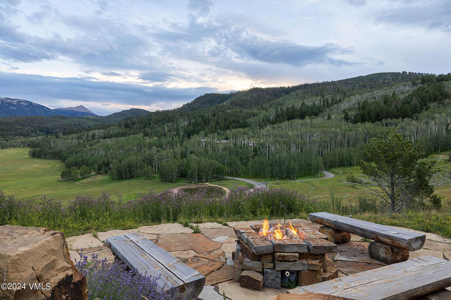 This image showcases a beautifully landscaped yard with a stone fire pit surrounded by wooden benches, creating an inviting outdoor living space. The backdrop features a scenic view of rolling hills, lush greenery, and a serene sky, enhancing the property's appeal and offering a tranquil atmosphere. The presence of wildflowers and mature trees adds to the natural beauty of the setting.
