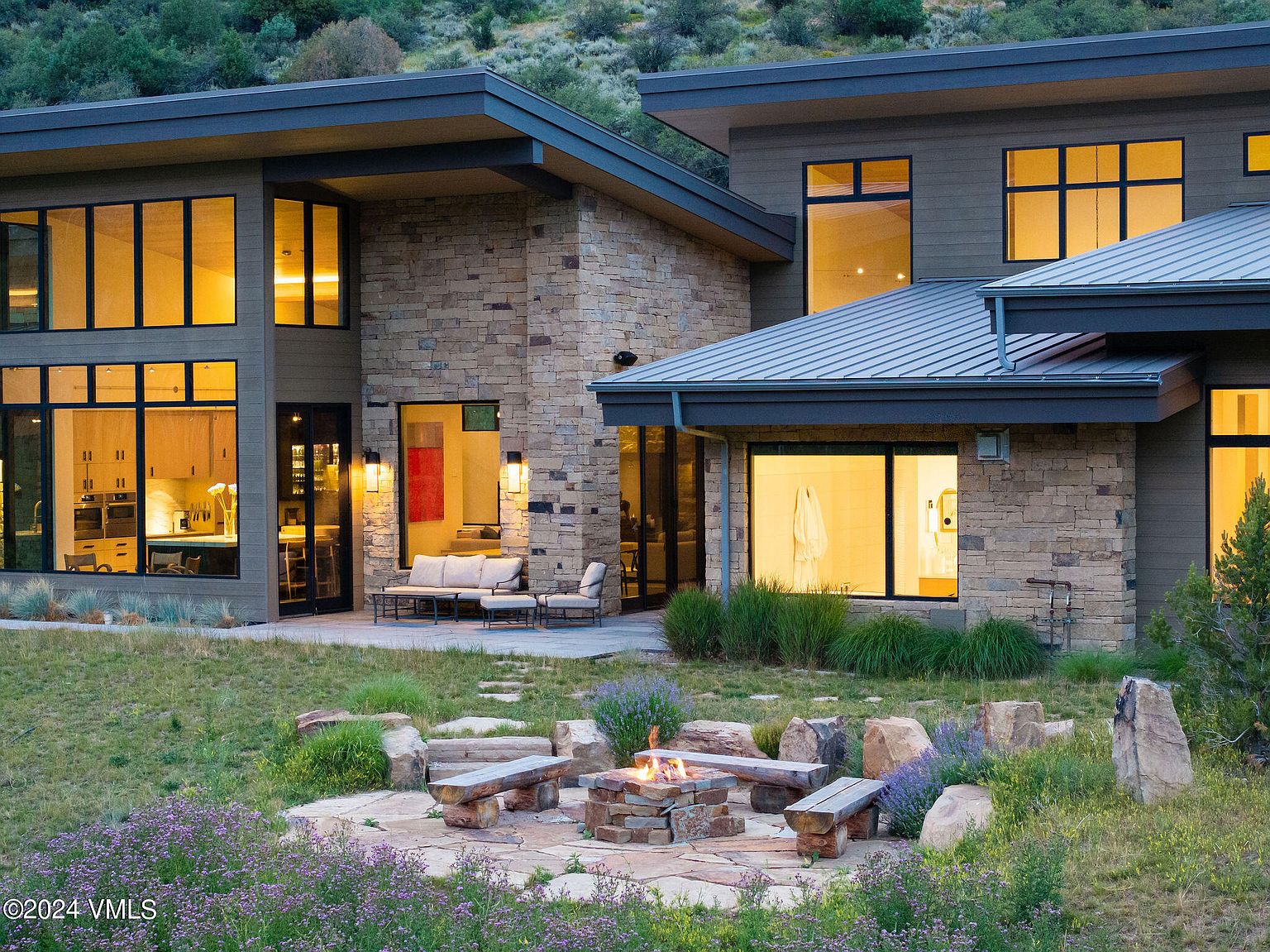 This image showcases the rear exterior of a modern home, featuring a stone facade, large windows providing ample natural light, and a cozy outdoor fire pit area with seating. The landscape is natural and blends seamlessly with the surrounding hillside, creating a serene and inviting atmosphere.
