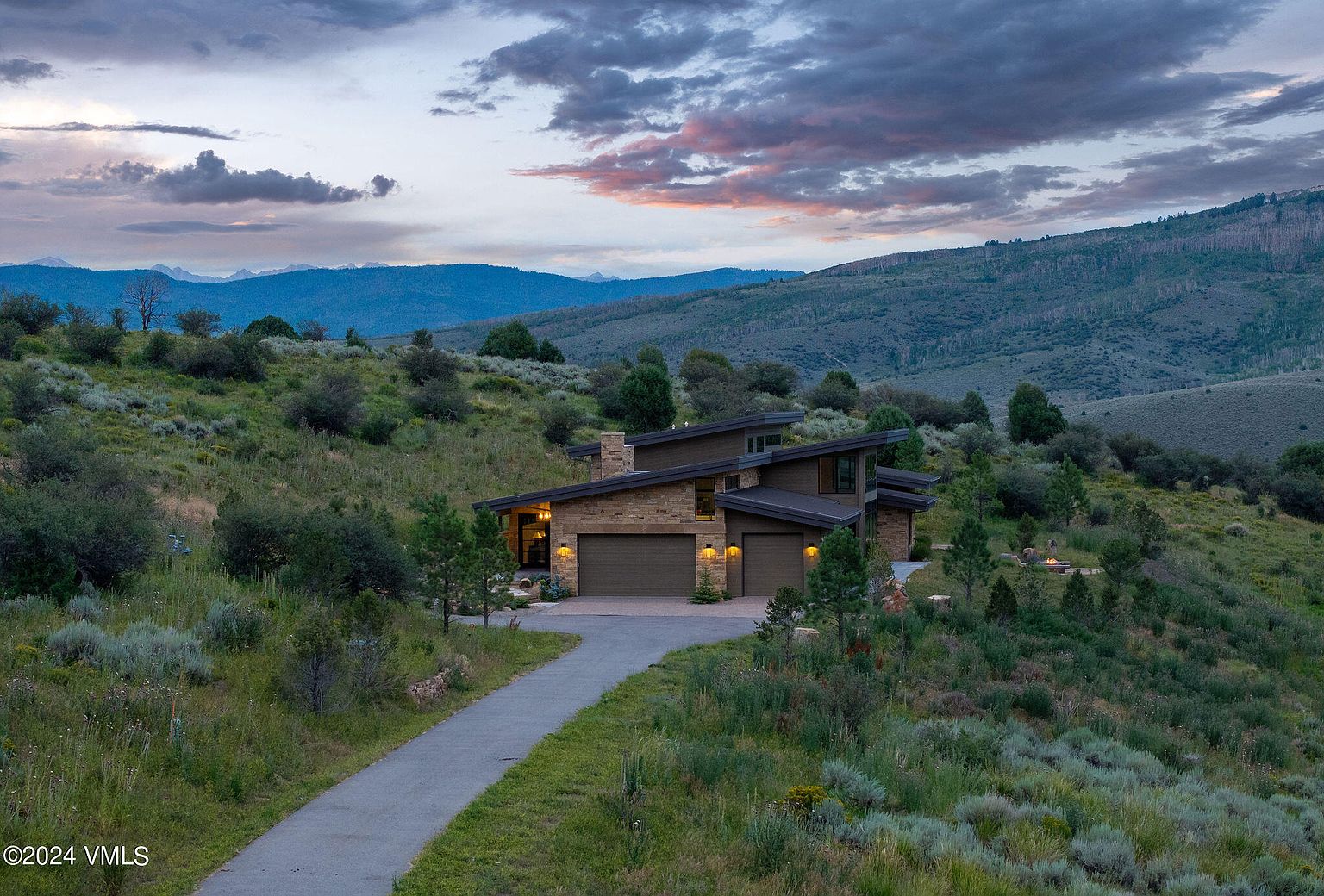 This image showcases the front exterior of a modern home nestled in a natural landscape. The house features a stone facade, a slanted roof, and a two-car garage. A paved driveway leads up to the property, surrounded by lush greenery and rolling hills, creating a sense of privacy and tranquility.