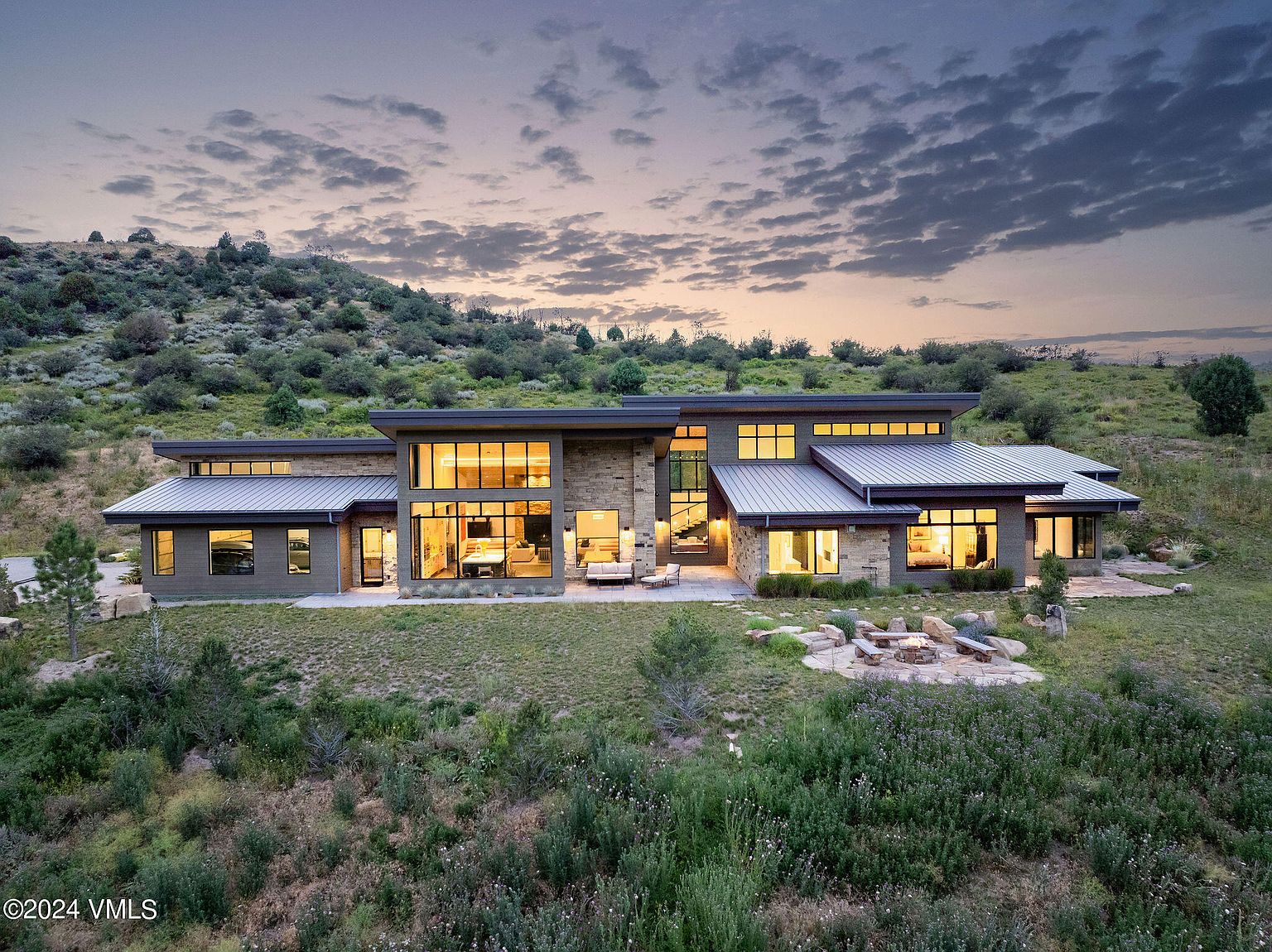 This image showcases the rear exterior of a modern home with large windows providing ample natural light. The property features a stone facade, a metal roof, and a well-maintained yard with a fire pit area. The surrounding landscape includes a hillside with natural vegetation, creating a sense of privacy and tranquility.