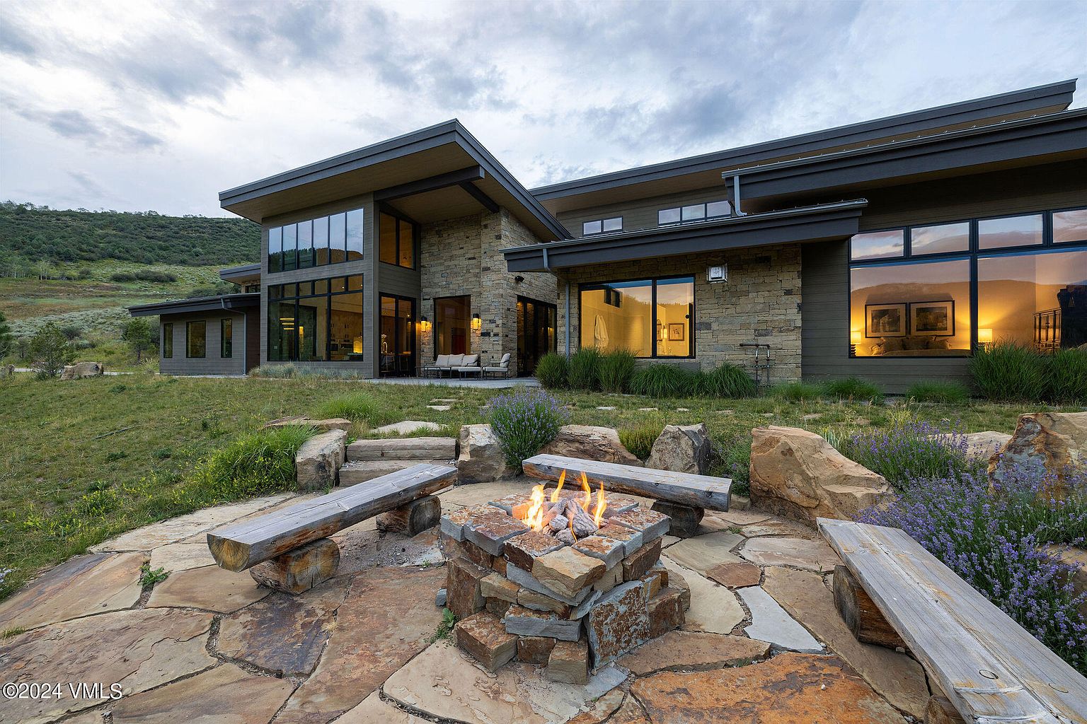 This image showcases the rear exterior of a modern home, featuring a stone facade, large windows, and a contemporary roofline. A stone fire pit with wooden benches creates an inviting outdoor living space, complemented by natural landscaping and a view of the surrounding hillside. The overall impression is one of luxury and tranquility.