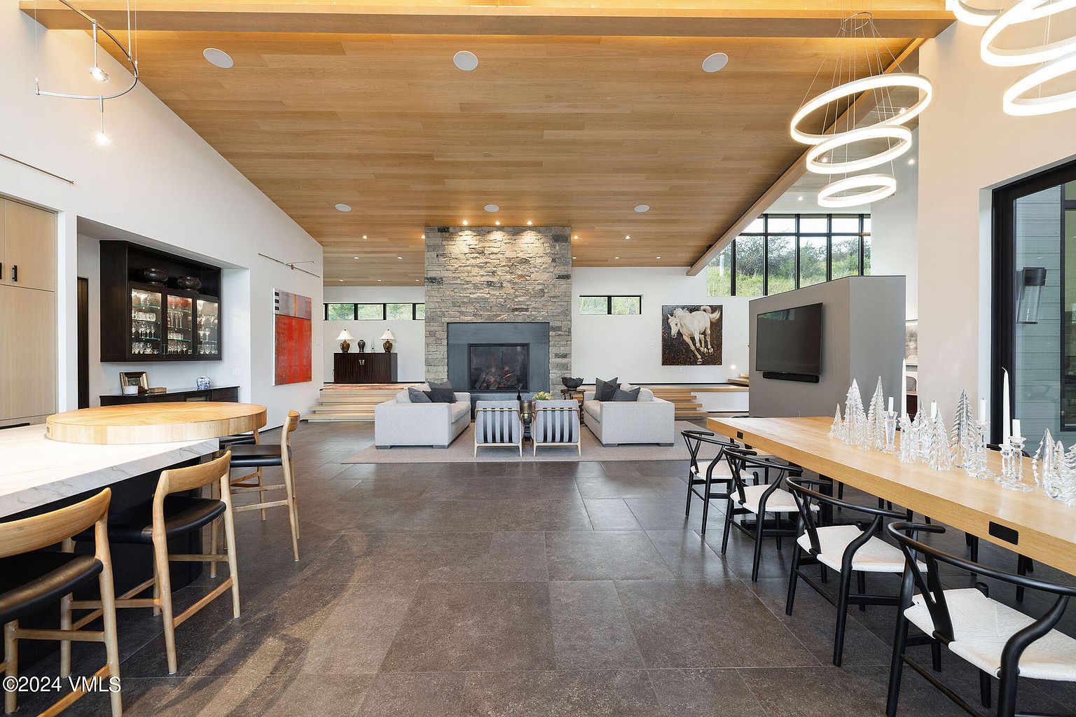 This expansive living room features a stone fireplace, modern furnishings, and a light wood ceiling. The open floor plan seamlessly connects the living area with the dining space, creating an inviting atmosphere. Large windows provide ample natural light, enhancing the contemporary design.