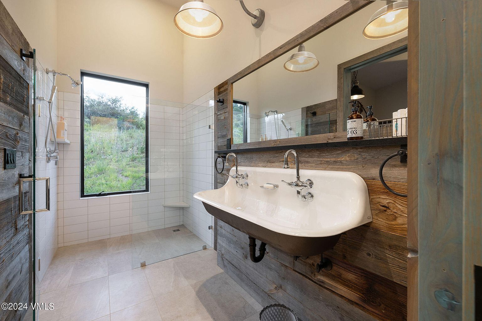 This primary bathroom features a unique trough sink with vintage-style faucets, set against a rustic wood-paneled wall. A glass-enclosed shower with white subway tile and a large window offering natural light complete the space. The design blends modern elements with rustic charm, creating a luxurious and inviting atmosphere.
