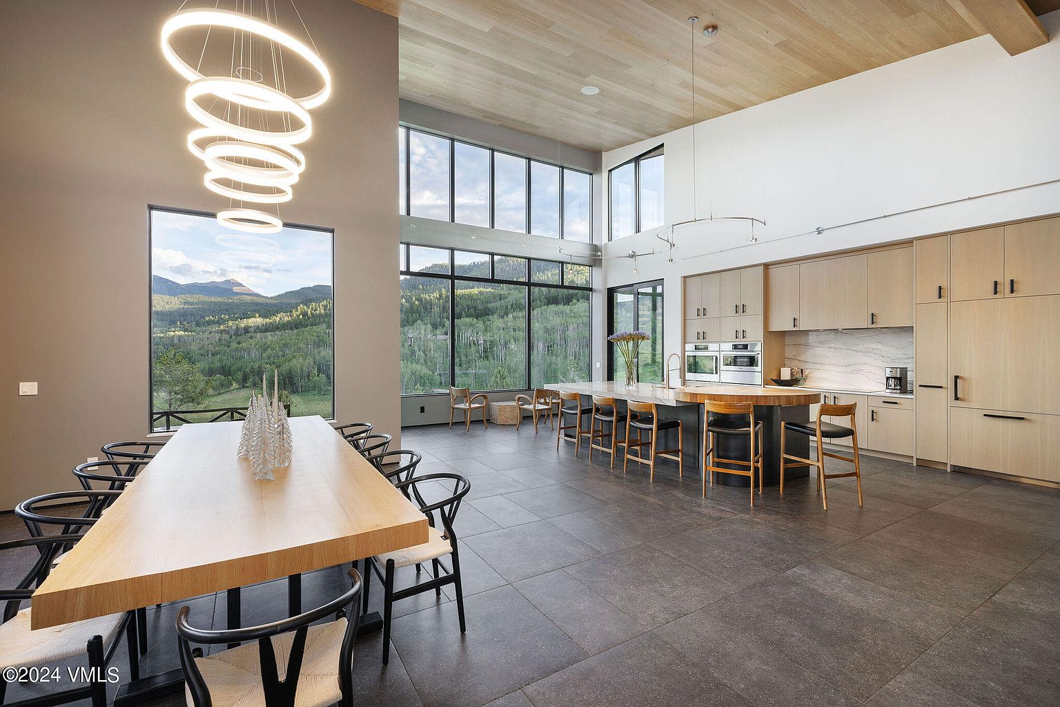 This interior shot showcases a modern kitchen and dining area with an open floor plan. The kitchen features light wood cabinetry, stainless steel appliances, and a large island with bar seating. The dining area includes a long wooden table with black chairs, illuminated by a unique circular chandelier, and large windows offer stunning views of the surrounding landscape.