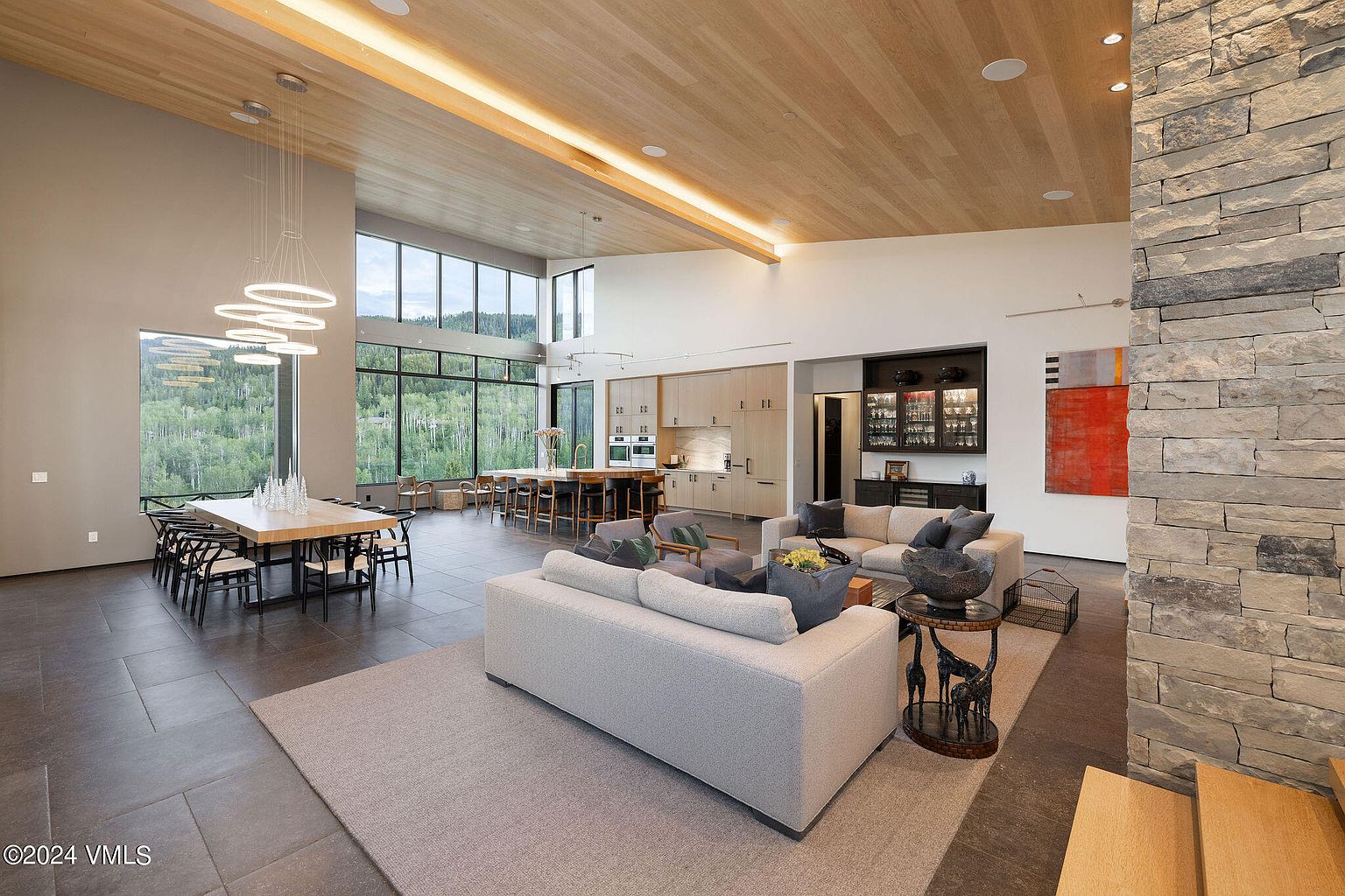 This expansive living room showcases a modern design with an open floor plan, seamlessly integrating the dining and kitchen areas. The space is characterized by high ceilings, large windows offering scenic views, and a sophisticated color palette. A stone accent wall adds texture and visual interest, enhancing the luxurious feel of the home.