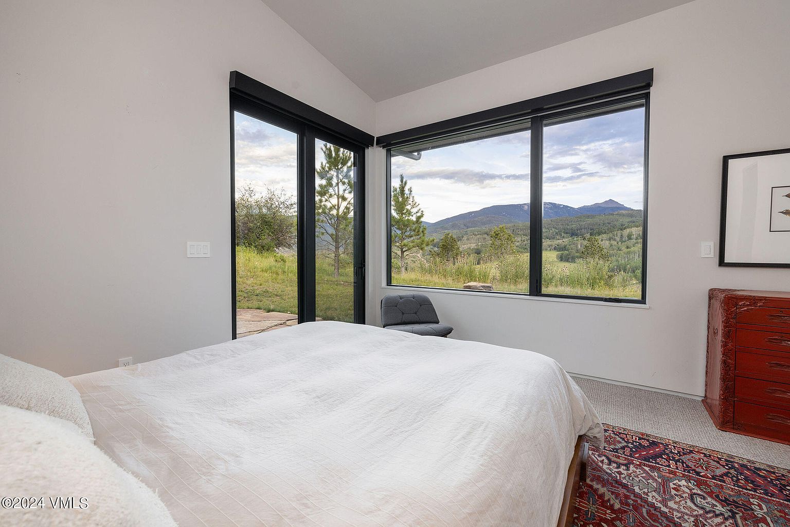 This primary bedroom features a large bed with white linens and expansive windows offering scenic views of the surrounding landscape. The room is minimalist in design with white walls, black window frames, and a touch of color from the rug and dresser, creating a serene and inviting atmosphere. The natural light and connection to the outdoors enhance the appeal of this space.