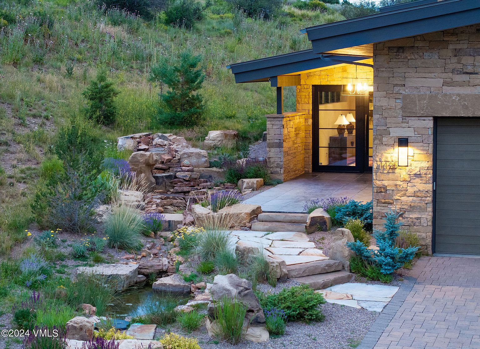 This image showcases the inviting entryway of a stone-clad home, featuring a beautifully landscaped front yard with a small waterfall and pond. Stone steps lead to the front door, which is framed by warm lighting and a modern design. The overall impression is one of natural elegance and curb appeal.
