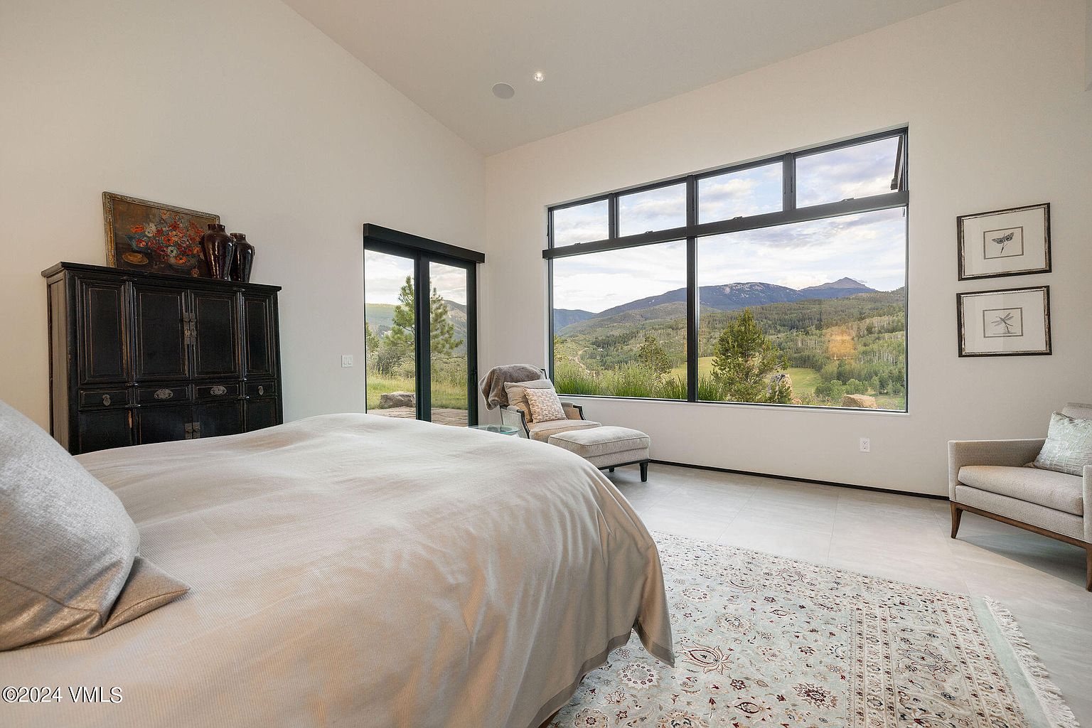 This primary bedroom features a large window with mountain views, a chaise lounge, and a neutral color palette. The room includes a dark wood armoire and a patterned area rug, creating a serene and luxurious atmosphere. The design emphasizes natural light and a connection to the outdoors.